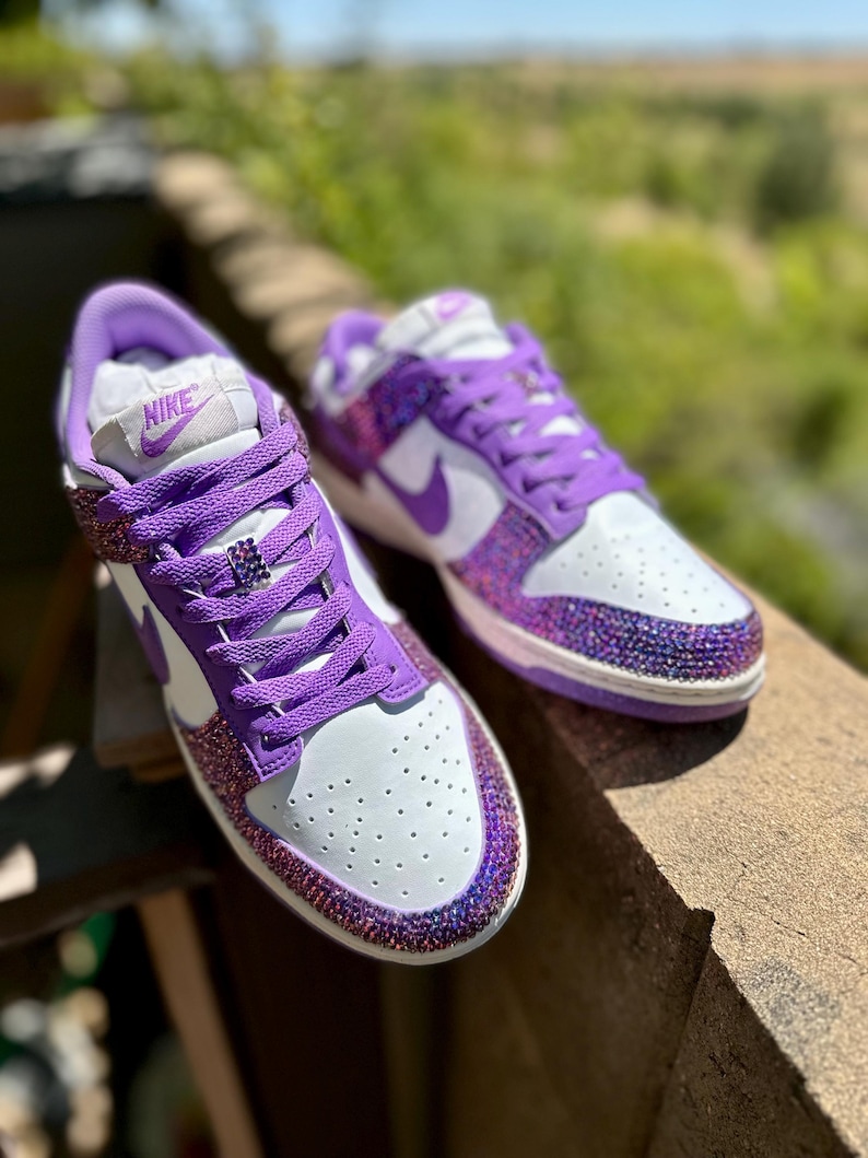 K&ouml;nnte beinhalten: Ein Paar wei&szlig;-lila Turnschuhe mit Strass an Zehen und Ferse. Die Turnschuhe haben lila Schn&uuml;rsenkel und ein lila Nike-Logo. Die Schuhe stehen auf einer Holzoberfl&auml;che.