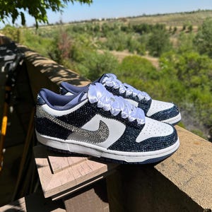 K&ouml;nnte beinhalten: Ein Paar marineblaue und wei&szlig;e Sneaker mit einem glitzernden Design. Die Schuhe haben einen wei&szlig;en Nike-Swoosh, wei&szlig;e Schn&uuml;rsenkel und eine wei&szlig;e Sohle. Die Sneaker sind mit kleinen, gl&auml;nzenden Verzierungen verziert.