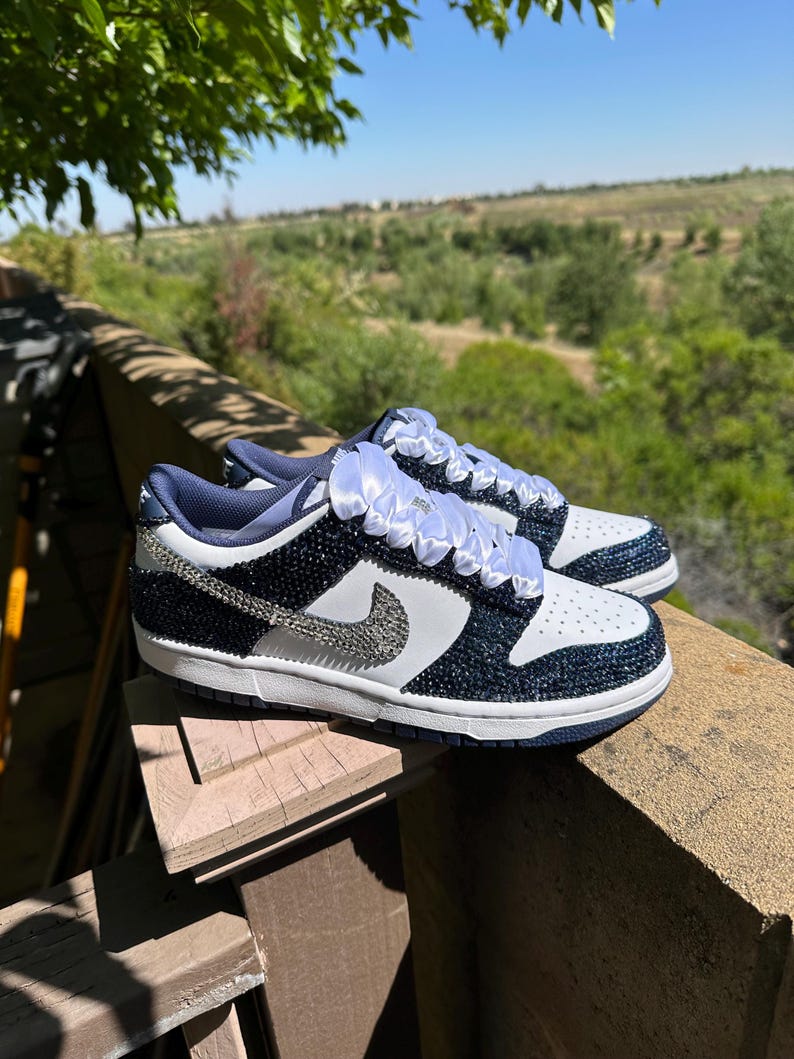 K&ouml;nnte beinhalten: Ein Paar marineblaue und wei&szlig;e Sneaker mit einem glitzernden Design. Die Schuhe haben einen wei&szlig;en Nike-Swoosh, wei&szlig;e Schn&uuml;rsenkel und eine wei&szlig;e Sohle. Die Sneaker sind mit kleinen, gl&auml;nzenden Verzierungen verziert.