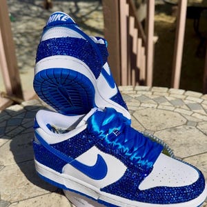 K&ouml;nnte beinhalten: Ein Paar ma&szlig;geschneiderte Nike Dunk Low Sneaker. Die Schuhe haben eine wei&szlig;e Lederbasis mit blauen Kristallverzierungen auf den Overlays und der Ferse. Der Nike Swoosh und die Schn&uuml;rsenkel sind blau. Auf der Zunge befindet sich das Nike-Logo.