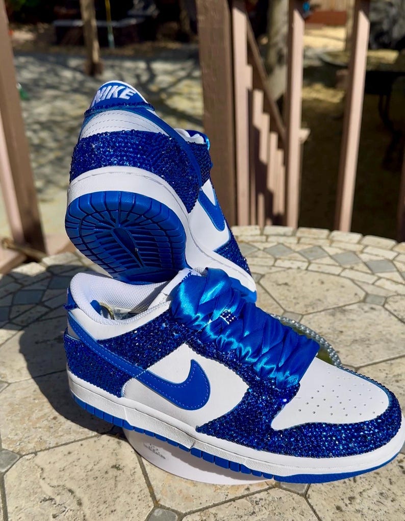 K&ouml;nnte beinhalten: Ein Paar ma&szlig;geschneiderte Nike Dunk Low Sneaker. Die Schuhe haben eine wei&szlig;e Lederbasis mit blauen Kristallverzierungen auf den Overlays und der Ferse. Der Nike Swoosh und die Schn&uuml;rsenkel sind blau. Auf der Zunge befindet sich das Nike-Logo.