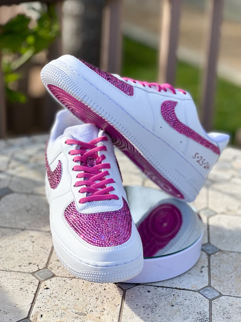 May include: White leather sneakers with pink glitter and a pink swoosh logo. The laces are pink and the shoes have a white sole.