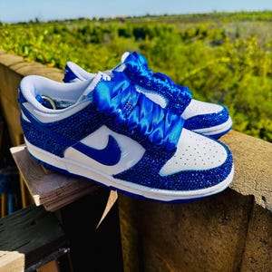 K&ouml;nnte beinhalten: Ein Paar blau-wei&szlig;e Sneaker mit einem Schmuckdesign. Die Schuhe haben einen blauen Nike-Swoosh und ein blaues Band an den Schn&uuml;rsenkeln. Die Sneaker werden im Freien pr&auml;sentiert.