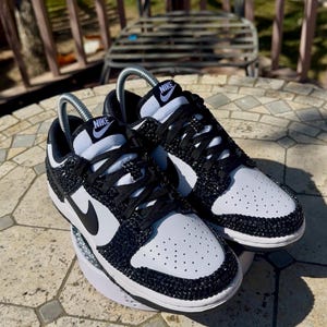 K&ouml;nnte beinhalten: Ein Paar schwarz-wei&szlig;e Nike Dunk Low Sneaker. Die Schuhe haben ein Obermaterial aus wei&szlig;em Leder mit schwarzen Akzenten, einschlie&szlig;lich des Nike-Swoosh und der Schn&uuml;rsenkel. Der Zehenbereich ist perforiert. Die Schuhe sind mit schwarzen Strasssteinen verziert.