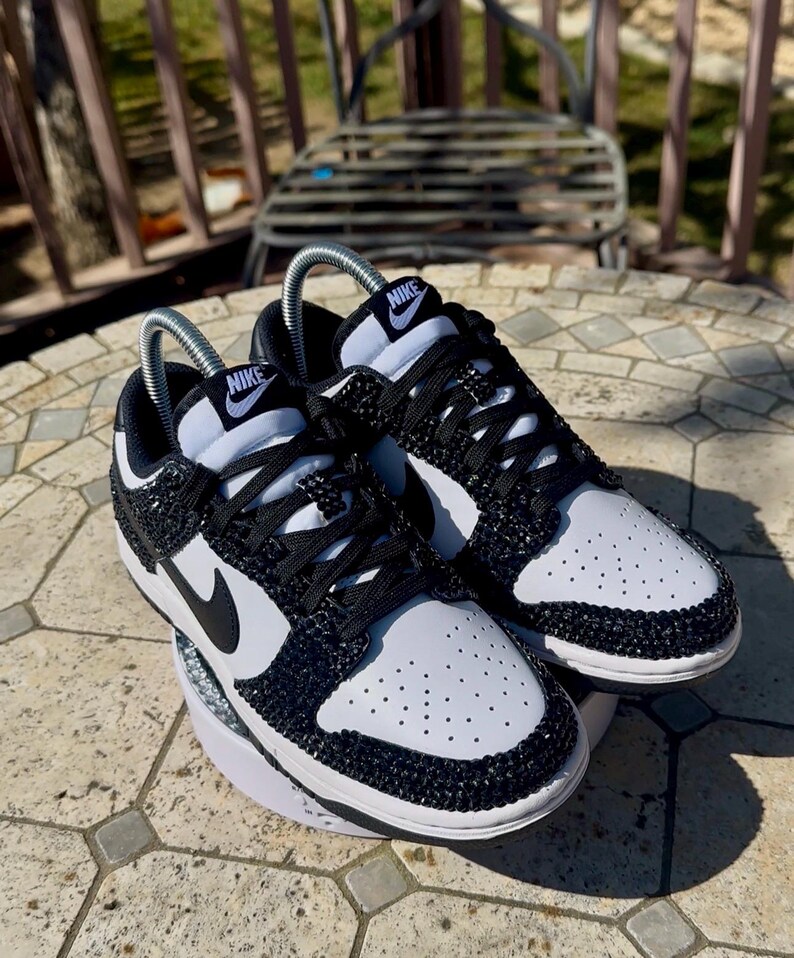 K&ouml;nnte beinhalten: Ein Paar schwarz-wei&szlig;e Nike Dunk Low Sneaker. Die Schuhe haben ein Obermaterial aus wei&szlig;em Leder mit schwarzen Akzenten, einschlie&szlig;lich des Nike-Swoosh und der Schn&uuml;rsenkel. Der Zehenbereich ist perforiert. Die Schuhe sind mit schwarzen Strasssteinen verziert.