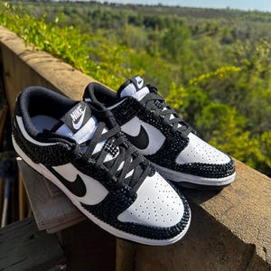 K&ouml;nnte beinhalten: Ein Paar wei&szlig;-schwarze Nike Dunk Low Sneaker. Die Schuhe haben eine wei&szlig;e Basis mit schwarzen Akzenten, einschlie&szlig;lich des Nike-Swoosh und der Schn&uuml;rsenkel. Die schwarzen Bereiche sind mit kleinen, glitzernden schwarzen Kristallen verziert. Die Sneaker werden im Freien gezeigt.