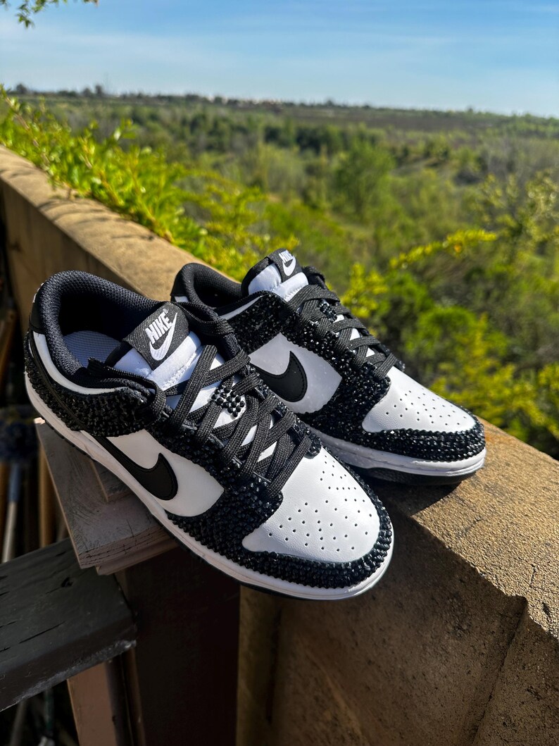 K&ouml;nnte beinhalten: Ein Paar wei&szlig;-schwarze Nike Dunk Low Sneaker. Die Schuhe haben eine wei&szlig;e Basis mit schwarzen Akzenten, einschlie&szlig;lich des Nike-Swoosh und der Schn&uuml;rsenkel. Die schwarzen Bereiche sind mit kleinen, glitzernden schwarzen Kristallen verziert. Die Sneaker werden im Freien gezeigt.