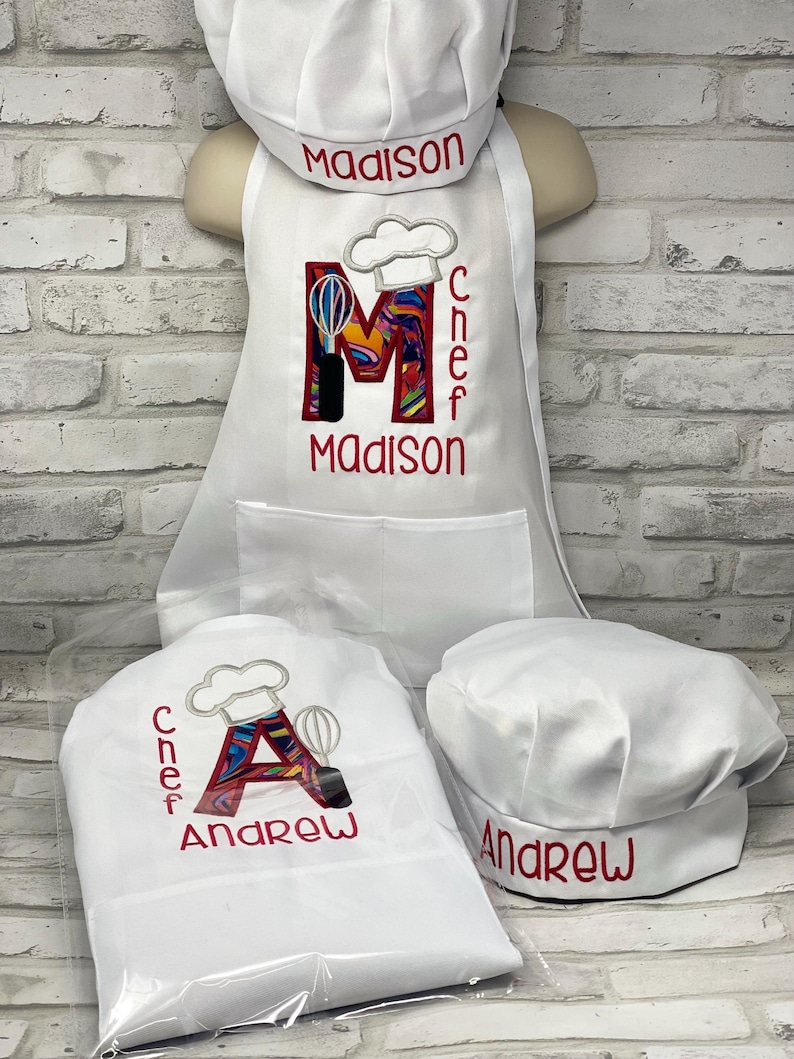 K&ouml;nnte beinhalten: Wei&szlig;es Kinderkochset mit Sch&uuml;rze, Kochm&uuml;tze und Kochhut. Die Sch&uuml;rze zeigt einen gro&szlig;en Buchstaben "M" mit farbenfrohem Design, das Wort "CHEF" und den Namen "Madison" in Rot. Der Kochhut hat auch den Namen "Madison" in Rot gestickt.