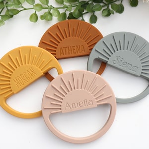 May include: Four silicone teethers shaped like suns with a personalized name on each. The teethers are in the colors mustard yellow, burnt orange, light pink, and gray.