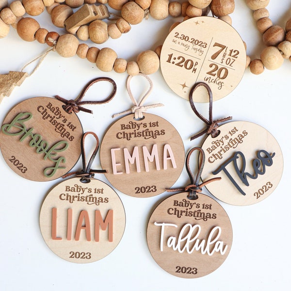 Puede incluir: Cinco adornos redondos de madera con el texto "Baby's 1st Christmas" y un nombre en cada adorno. Los nombres son Brook, Emma, Theo, Liam y Tallula. Los adornos están decorados con diferentes colores y fuentes. Los adornos cuelgan de cordones de cuero marrón.