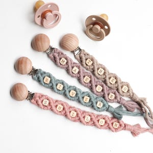 May include: Three macrame pacifier clips featuring wooden beads with the letters M-A-E-V-E, R-I-V-E-R, and S-H-I-L-O-H, each attached to a round wooden clip. Two silicone pacifiers in muted pink and beige tones are positioned above the clips.