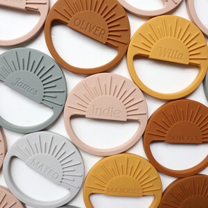 May include: A collection of personalized silicone teethers in various colors, each featuring a sun design and a name. The teethers are in shades of brown, gray, pink, and yellow.