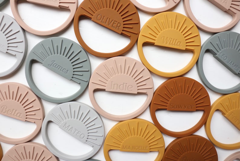 May include: A collection of personalized silicone teethers in various colors, each featuring a sun design and a name. The teethers are in shades of brown, gray, pink, and yellow.