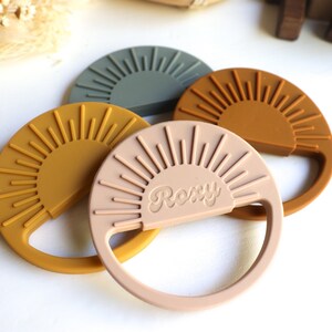 May include: Three silicone teething toys shaped like suns with a raised sunburst design. The toys are in shades of green, brown, and pink. The pink toy has the word "Roxy" embossed on it.