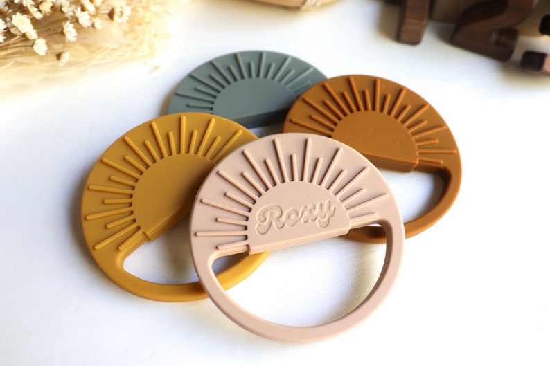 May include: Three silicone teething toys shaped like suns with a raised sunburst design. The toys are in shades of green, brown, and pink. The pink toy has the word "Roxy" embossed on it.