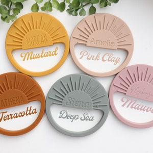 May include: Five silicone teething rings in different colors with a sunburst design and a name and color written on each. The colors are mustard, pink clay, terracotta, deep sea, and mauve.
