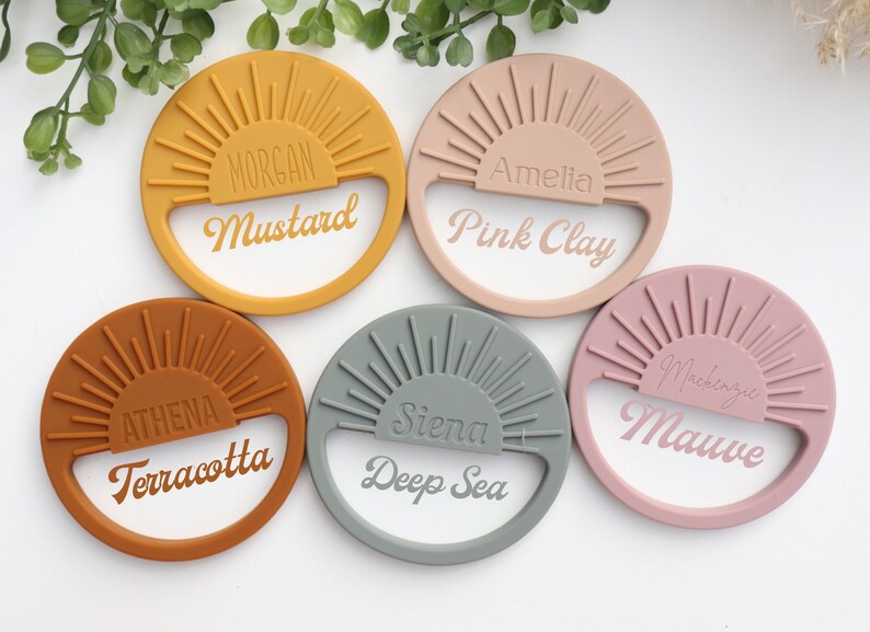 May include: Five silicone teething rings in different colors with a sunburst design and a name and color written on each. The colors are mustard, pink clay, terracotta, deep sea, and mauve.