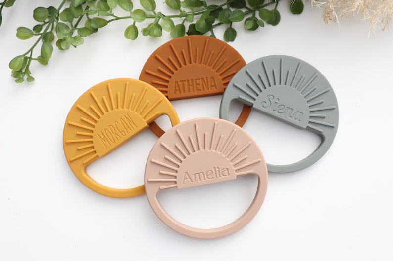May include: Four silicone teethers shaped like suns with a personalized name on each. The teethers are in the colors mustard yellow, burnt orange, light pink, and gray.