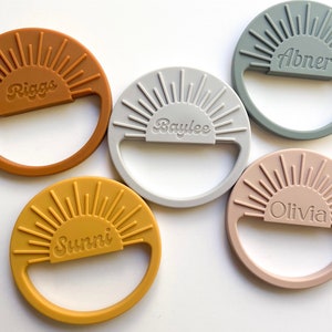 May include: Five silicone teething rings in different colors with a sunburst design and personalized names: Riggs, Baylee, Abner, Olivia, and Sunni.