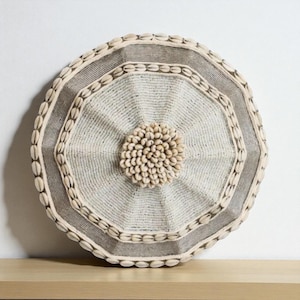 May include: A round, woven wall decor piece with a central cluster of small, beige shells. The design features concentric octagonal and circular patterns, with a border of small shells and a striped outer edge. The color palette is neutral, with beige and gray tones.