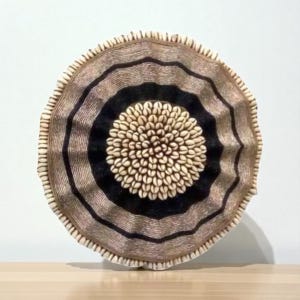 May include: Round wall art featuring a central cluster of seashells surrounded by concentric rings of black, tan, and beige. The outer edge has a textured, ruffled appearance. This decorative piece adds a natural, coastal theme to any space.