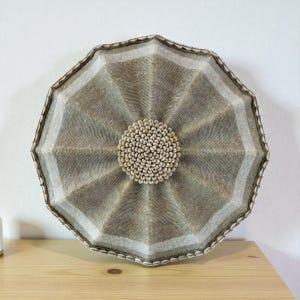 May include: A decorative wall hanging with a unique design. The piece features a central circular element surrounded by a radiating pattern in shades of gray and beige. The outer edge is a darker brown, creating a striking contrast. The central circle is made of small, light-colored beads.