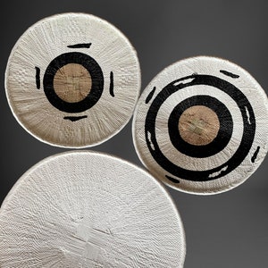 May include: Three woven, circular decorative pieces. The pieces are primarily white with black and tan circular designs. The designs vary, with one featuring concentric black rings and another with a black circle and accents. The third piece is mostly white.