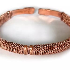 Pode incluir: Um bracelete de cobre com um design tecido e quatro acentos redondos. O bracelete é aberto e ajustável.