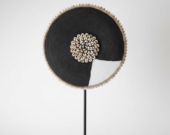 May include: A circular decorative object on a black metal stand. The disc is covered in black beads with a white beaded section, a central cluster of white cowrie shells, and a border of small shells along the outer edge of the circular frame.