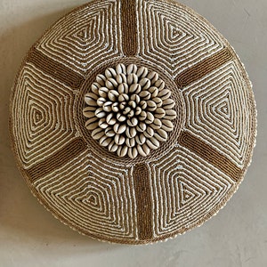Puede incluir: Un cojín redondo de cuentas con un patrón geométrico en dorado y blanco. El centro del cojín presenta un diseño circular hecho de conchas blancas.