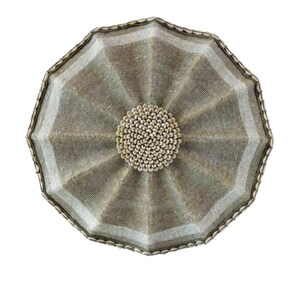May include: A decorative, round, woven wall hanging with a central circular design. The piece features a radial pattern of alternating light and dark beige segments, with a beaded center and edge.
