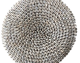May include: A circular wall hanging composed of numerous small, off-white cowrie shells arranged in a dense, concentric pattern. The shells are tightly packed, radiating from the center toward the outer edge, creating a textured, layered appearance.