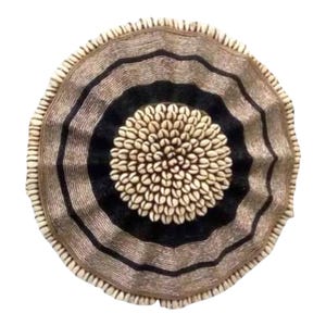 May include: Circular wall decor featuring concentric rings of black and tan, adorned with a central cluster of white seashells. The outer edge is trimmed with small, cream-colored shells, creating a textured, decorative piece.