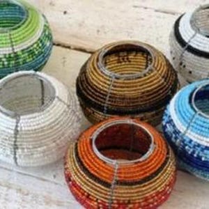 Könnte beinhalten: Eine Sammlung von sieben Kerzenhaltern aus Perlen in verschiedenen Farben. Die runden Halter haben offene Oberteile und sind mit farbigen Perlen in Blau-, Grün-, Orange-, Braun- und Weißtönen verziert. Sie sind auf einer hellen Holzoberfläche angeordnet.
