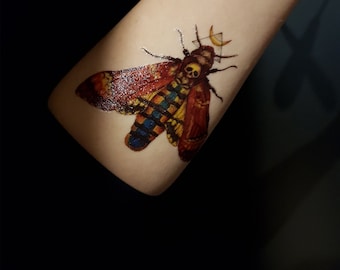 May include: A temporary tattoo of a colorful moth with a skull on its head. The moth is surrounded by a geometric design with a crescent moon.