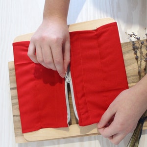 以下が含まれることがあります： 木製の台座に置かれた、赤い布製のアイテム。銀色のジッパーが付いており、手で開けられています。感覚玩具または教育玩具のように見えます。