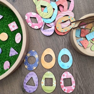 May include: Wooden Easter-themed educational toy set. Includes colorful egg-shaped cutouts with geometric shapes and patterns, along with a wooden bowl filled with smaller shapes and a tray with green faux grass.