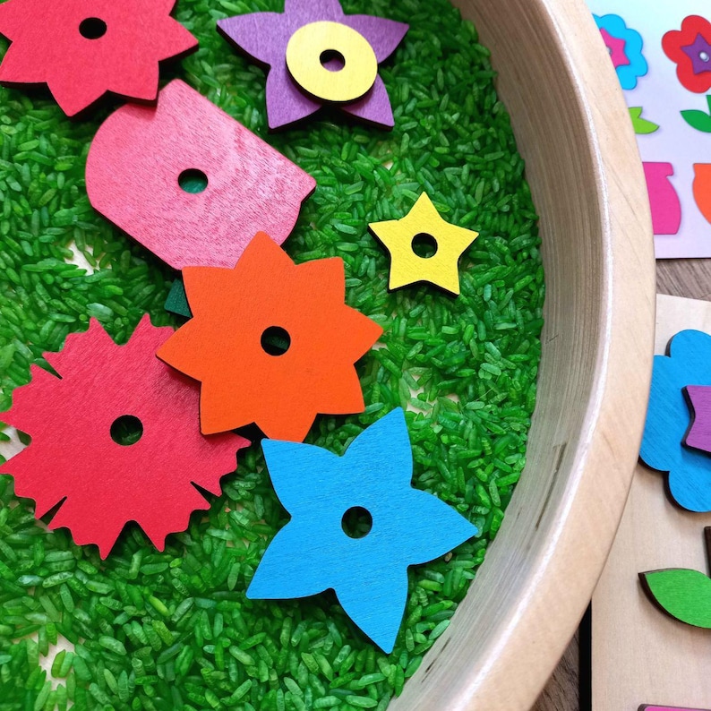 May include: A wooden tray filled with green rice and colorful wooden flower shapes. The flowers have a hole in the center and are red, orange, blue, purple, and yellow.