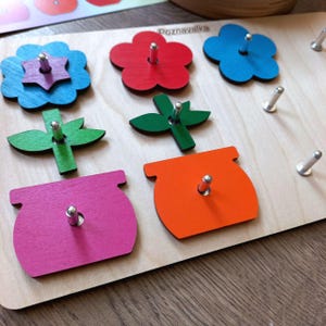 May include: A wooden educational toy with colorful shapes and pegs. The shapes include a flower, a pot, and a leaf. The toy is labeled "Poznavalka" which is Polish for "learning".
