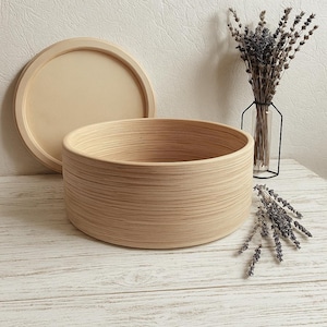 May include: A light brown, round wooden bowl with a matching lid. The bowl has a textured, striped pattern. A vase with dried lavender and a sprig of lavender are also in the image.