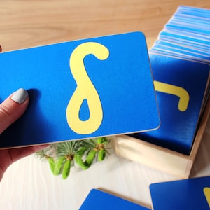 May include: A set of blue wooden cards with yellow felt letters. The cards are arranged in a wooden box. The letter on the top card is the letter "S".