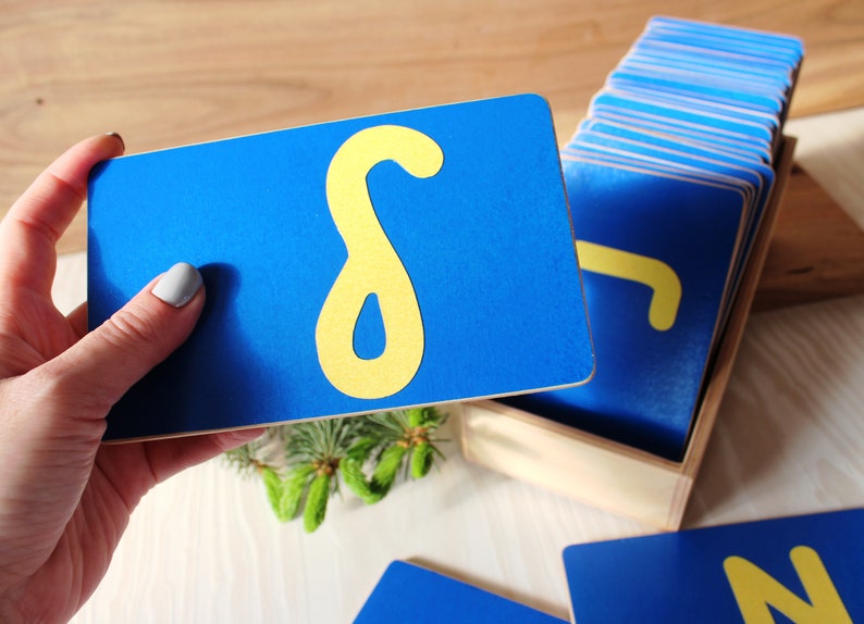 May include: A set of blue wooden cards with yellow felt letters. The cards are arranged in a wooden box. The letter on the top card is the letter "S".