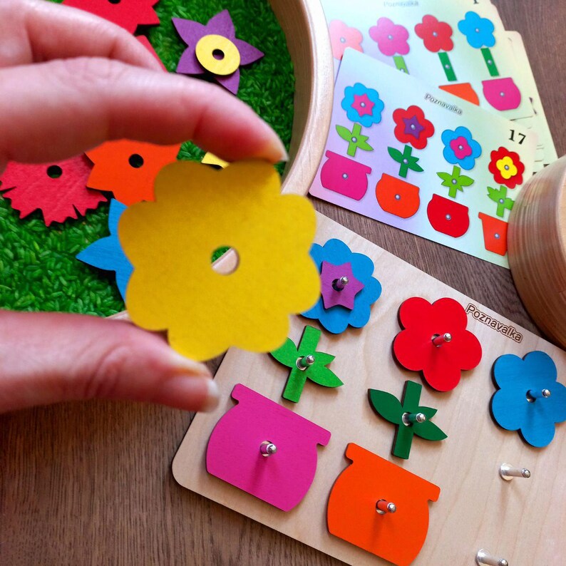May include: A wooden educational toy with a flower-shaped puzzle piece in a hand. The puzzle piece is yellow and has a hole in the center. The toy has a wooden base with different colored flowers and pots.