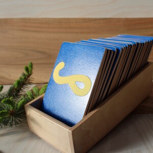 May include: A wooden box filled with blue cards, each with a yellow letter or shape on it. The box is sitting on a wooden surface.