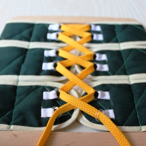 Puede incluir: Un tablero de lazado de tela verde y blanco con cordones amarillos. Los cordones se enhebran a trav&eacute;s de los agujeros de la tela y se atan en un patr&oacute;n de cruz.