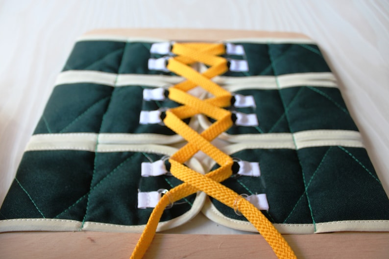 Puede incluir: Un tablero de lazado de tela verde y blanco con cordones amarillos. Los cordones se enhebran a trav&eacute;s de los agujeros de la tela y se atan en un patr&oacute;n de cruz.