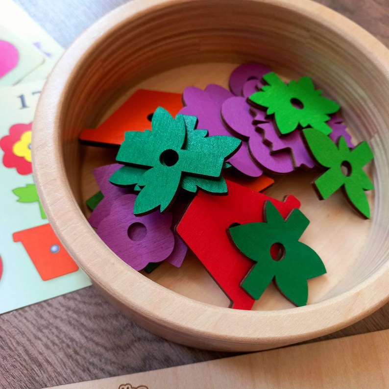 May include: A wooden bowl filled with colorful wooden flower shapes. The shapes are in shades of red, green, purple, and yellow. The bowl is made of light wood and has a natural finish.