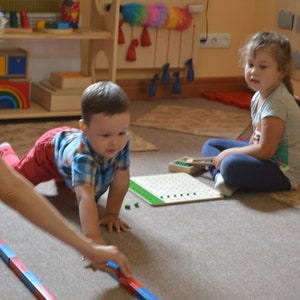 Pu&ograve; includere: Due bambini giocano con giocattoli educativi in ​​un'aula. Un bambino piccolo striscia sul pavimento mentre una bambina piccola &egrave; seduta sul pavimento. Il bambino sta giocando con un set di bastoncini di legno rossi e blu. La bambina sta giocando con una tavola di legno con pioli verdi.