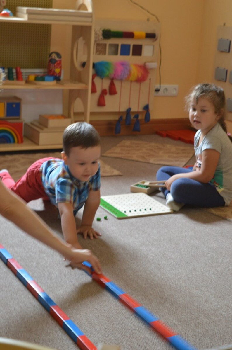 Pu&ograve; includere: Due bambini giocano con giocattoli educativi in ​​un'aula. Un bambino piccolo striscia sul pavimento mentre una bambina piccola &egrave; seduta sul pavimento. Il bambino sta giocando con un set di bastoncini di legno rossi e blu. La bambina sta giocando con una tavola di legno con pioli verdi.