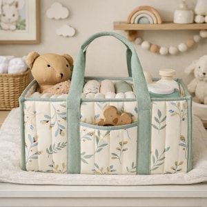 May include: A quilted baby caddy with a floral pattern in cream, blue, and yellow. The caddy has mint green handles and trim, and a front pocket with a wooden toy. Inside are baby essentials, including a teddy bear and wipes.